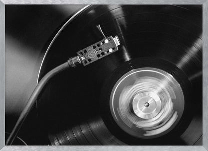 A framed, black and white, close-up photograph of a record player in action. The turntable is spinning a vinyl record, creating a motion blur on the center label. The tonearm and stylus are positioned over the grooved surface of the record. Poster