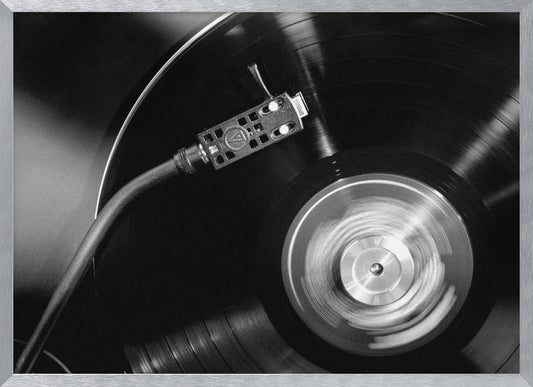 A framed, black and white, close-up photograph of a record player in action. The turntable is spinning a vinyl record, creating a motion blur on the center label. The tonearm and stylus are positioned over the grooved surface of the record. Poster