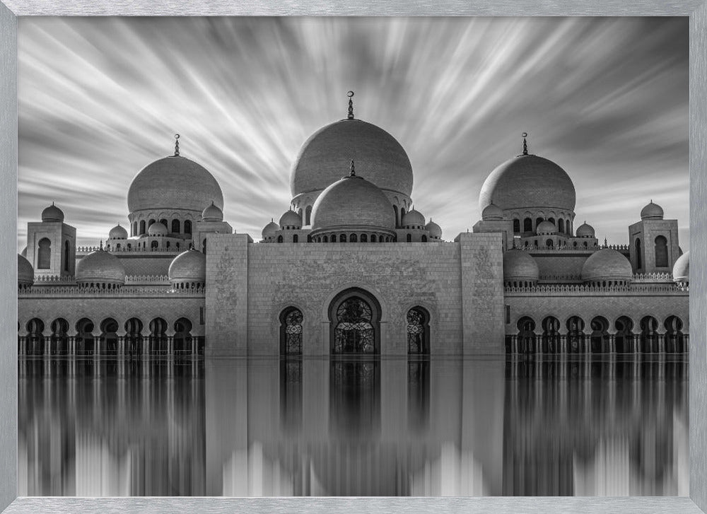 A stunning black and white long-exposure photograph of a grand mosque with multiple domes, reflected perfectly in calm water. The sky above is dramatic, with clouds streaking outwards, and the image is presented in a silver frame. Decor