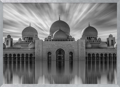 A stunning black and white long-exposure photograph of a grand mosque with multiple domes, reflected perfectly in calm water. The sky above is dramatic, with clouds streaking outwards, and the image is presented in a silver frame. Decor