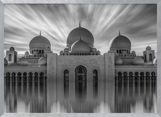 A stunning black and white long-exposure photograph of a grand mosque with multiple domes, reflected perfectly in calm water. The sky above is dramatic, with clouds streaking outwards, and the image is presented in a silver frame. Decor