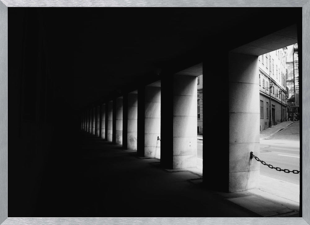 A high-contrast, black and white photograph from the perspective of a dark, covered walkway lined with a row of thick pillars. Light from an adjacent street highlights the pillars, creating a dramatic play of light and shadow and a strong sense of depth. Print