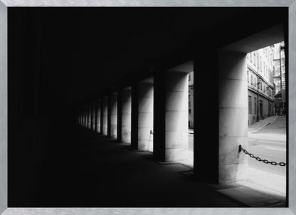 A high-contrast, black and white photograph from the perspective of a dark, covered walkway lined with a row of thick pillars. Light from an adjacent street highlights the pillars, creating a dramatic play of light and shadow and a strong sense of depth. Print
