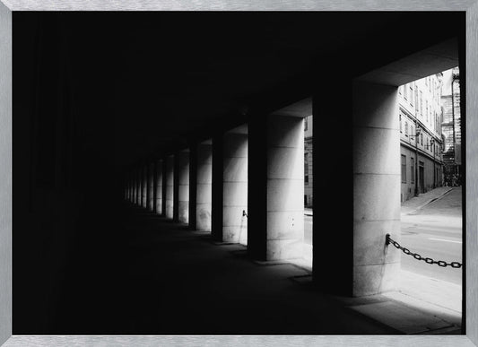 A high-contrast, black and white photograph from the perspective of a dark, covered walkway lined with a row of thick pillars. Light from an adjacent street highlights the pillars, creating a dramatic play of light and shadow and a strong sense of depth. Print