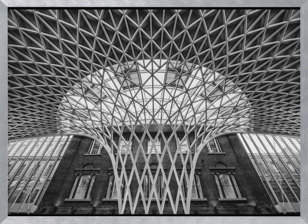 A framed, black and white photograph of the Western Concourse at King's Cross Station, London. The view is from a low angle, looking up at the intricate, white, funnel-like lattice roof structure as it spreads out across the ceiling, contrasting with the classic brick facade of the building behind it. Decor