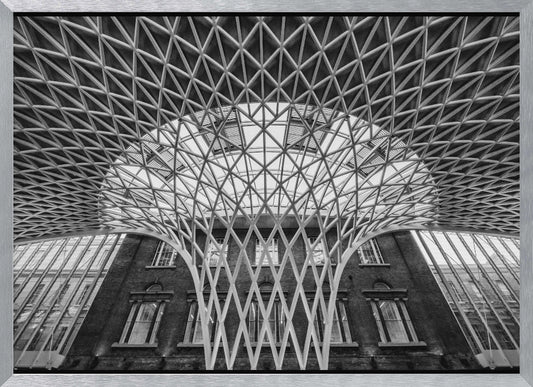 A framed, black and white photograph of the Western Concourse at King's Cross Station, London. The view is from a low angle, looking up at the intricate, white, funnel-like lattice roof structure as it spreads out across the ceiling, contrasting with the classic brick facade of the building behind it. Decor