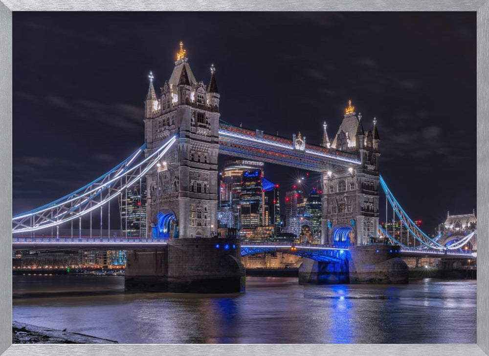 A vibrant night photograph of the iconic Tower Bridge in London, illuminated with bright white and electric blue lights. The dark River Thames in the foreground reflects the bridge and the lit-up modern city skyline in the background, all under a dark, cloudy sky. The image is enclosed in a silver frame. Decor