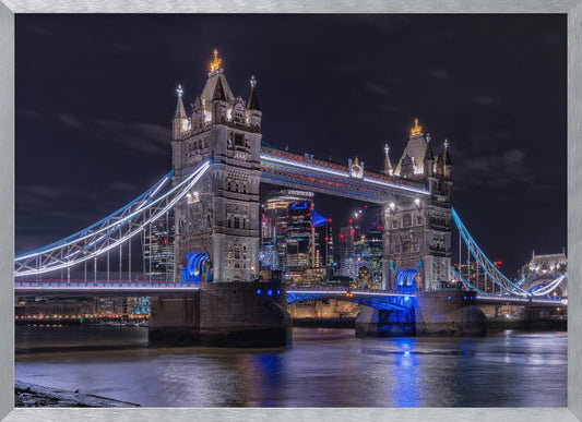 A vibrant night photograph of the iconic Tower Bridge in London, illuminated with bright white and electric blue lights. The dark River Thames in the foreground reflects the bridge and the lit-up modern city skyline in the background, all under a dark, cloudy sky. The image is enclosed in a silver frame. Decor