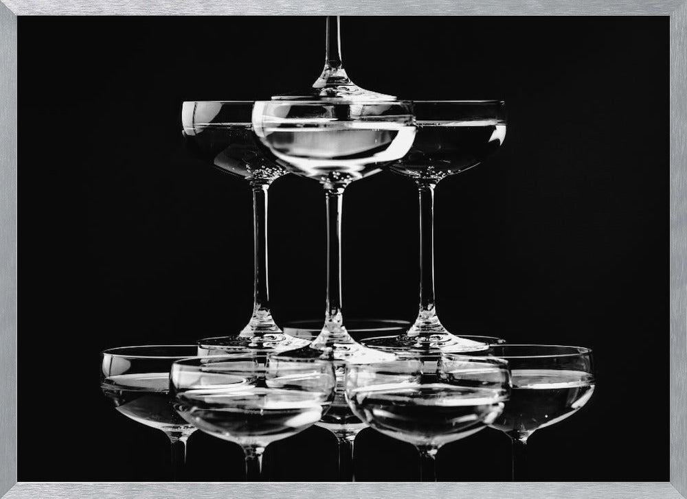 A black and white photograph of a champagne tower made of coupe glasses stacked in a pyramid shape against a solid black background, all within a silver frame. Print
