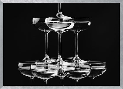 A black and white photograph of a champagne tower made of coupe glasses stacked in a pyramid shape against a solid black background, all within a silver frame. Print