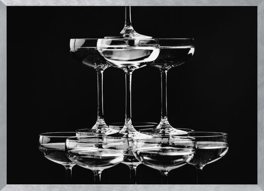 A black and white photograph of a champagne tower made of coupe glasses stacked in a pyramid shape against a solid black background, all within a silver frame. Print
