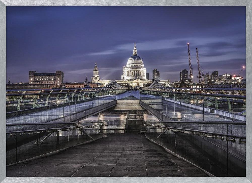 A nighttime photograph from the Millennium Bridge leading towards the brightly illuminated St. Paul's Cathedral against a dark blue twilight sky in London, all enclosed in a silver frame. Poster