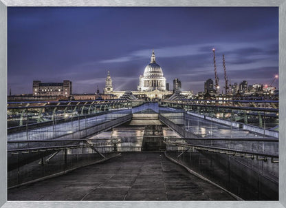 A nighttime photograph from the Millennium Bridge leading towards the brightly illuminated St. Paul's Cathedral against a dark blue twilight sky in London, all enclosed in a silver frame. Poster
