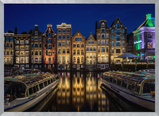 A vibrant night-time photograph of a canal in Amsterdam, flanked by two tour boats in the foreground. Traditional Dutch canal houses are brightly lit, casting warm yellow reflections on the dark water under a deep blue sky. Print