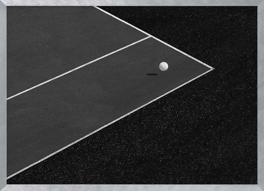 A minimalist black and white overhead photograph of a white ball resting in the corner of a dark tennis or paddle court. The composition focuses on the strong geometric angles created by the white boundary lines against the dark, textured asphalt, all within a silver frame. Artwork