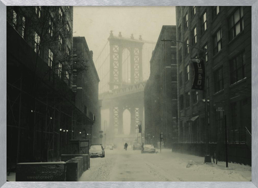 A framed, vintage-style black and white photograph capturing a snowy day in DUMBO, Brooklyn. The view looks down a snow-covered street lined with dark buildings, perfectly framing the Manhattan Bridge in the foggy distance. A few cars are parked along the street, and snow is visibly falling, creating a quiet, atmospheric winter scene. Decor