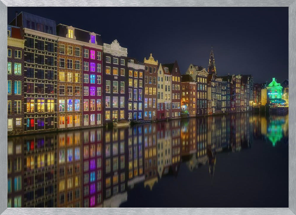 A vibrant nighttime photograph of colorful canal houses in Amsterdam, with their glowing lights beautifully reflected in the still water of the canal below, all framed in silver. Print
