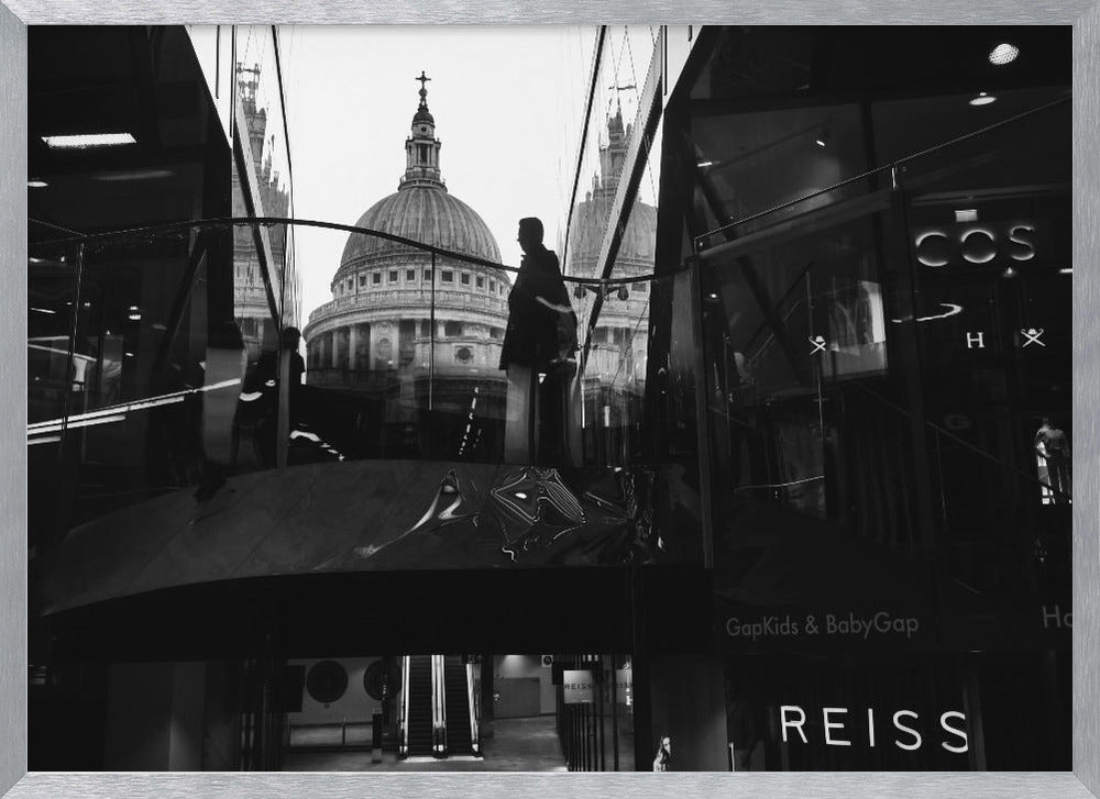 A framed, black and white photograph capturing the reflection of St. Paul's Cathedral in the glass of a modern shopping center. A person's silhouette stands on an interior balcony, juxtaposed against the historic dome, while the mall's interior and store signs are visible in the foreground. Print