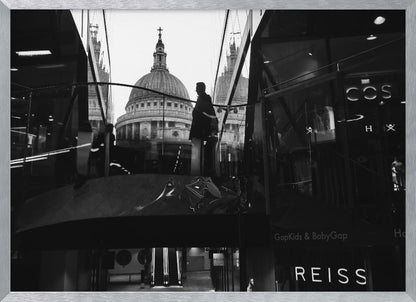 A framed, black and white photograph capturing the reflection of St. Paul's Cathedral in the glass of a modern shopping center. A person's silhouette stands on an interior balcony, juxtaposed against the historic dome, while the mall's interior and store signs are visible in the foreground. Print
