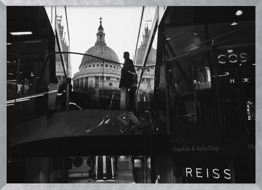 A framed, black and white photograph capturing the reflection of St. Paul's Cathedral in the glass of a modern shopping center. A person's silhouette stands on an interior balcony, juxtaposed against the historic dome, while the mall's interior and store signs are visible in the foreground. Print