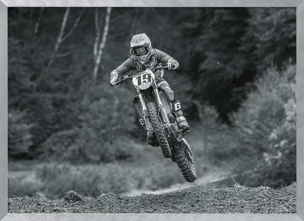 A dynamic black and white photograph of a motocross racer in mid-air, jumping over a dirt mound. The rider, wearing full protective gear and a helmet, is on a dirt bike with the number 19 plate. The background is a soft-focus forest, and the entire image is enclosed in a silver frame. Poster