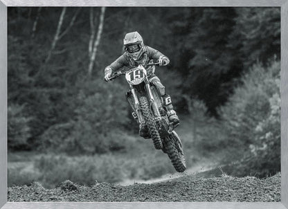 A dynamic black and white photograph of a motocross racer in mid-air, jumping over a dirt mound. The rider, wearing full protective gear and a helmet, is on a dirt bike with the number 19 plate. The background is a soft-focus forest, and the entire image is enclosed in a silver frame. Poster