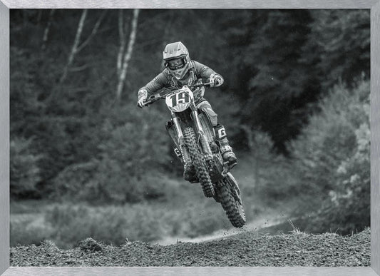 A dynamic black and white photograph of a motocross racer in mid-air, jumping over a dirt mound. The rider, wearing full protective gear and a helmet, is on a dirt bike with the number 19 plate. The background is a soft-focus forest, and the entire image is enclosed in a silver frame. Poster