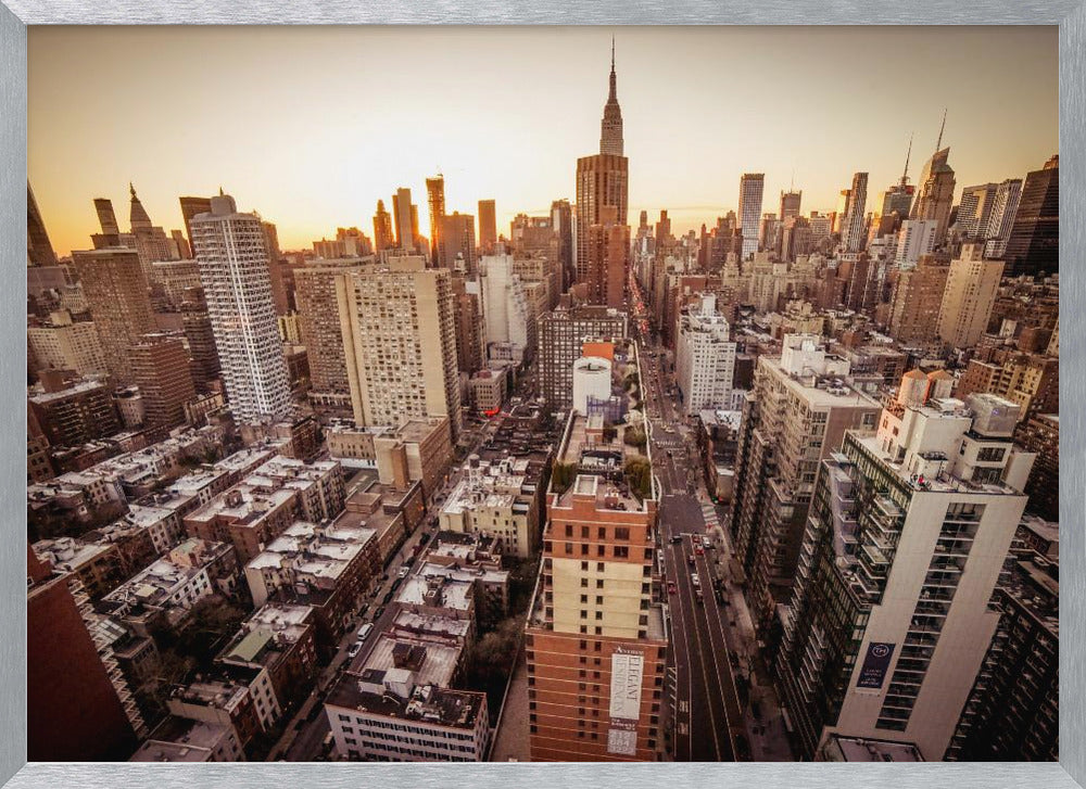 An aerial photograph of the New York City skyline at sunset, with the Empire State Building prominent against the glowing sky. The warm light of the setting sun bathes the dense urban landscape in a golden hue. The entire scene is enclosed in a silver, brushed metal frame. Print