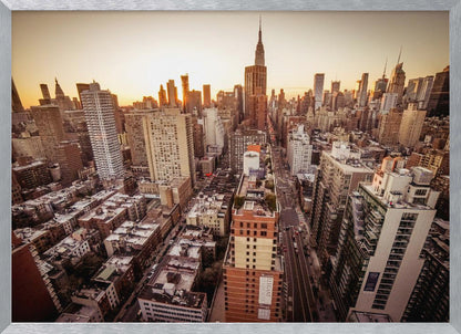 An aerial photograph of the New York City skyline at sunset, with the Empire State Building prominent against the glowing sky. The warm light of the setting sun bathes the dense urban landscape in a golden hue. The entire scene is enclosed in a silver, brushed metal frame. Print
