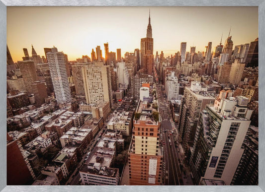 An aerial photograph of the New York City skyline at sunset, with the Empire State Building prominent against the glowing sky. The warm light of the setting sun bathes the dense urban landscape in a golden hue. The entire scene is enclosed in a silver, brushed metal frame. Print