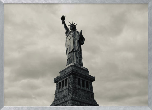 A low-angle, black and white photograph of the Statue of Liberty standing tall against a cloudy, dramatic sky, enclosed in a silver frame. Poster