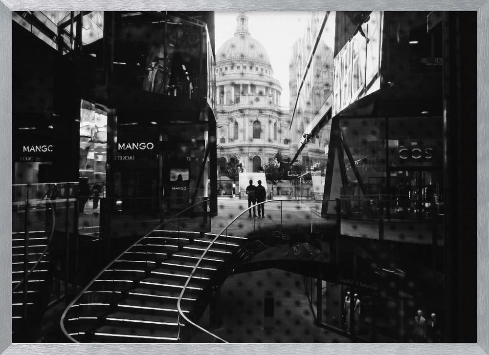 A framed, black and white photograph capturing a layered urban scene. The view, seen through a window with reflections of a curved, lit staircase, shows two people walking towards a large domed cathedral framed by modern glass buildings. Wall Art