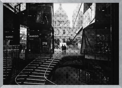 A framed, black and white photograph capturing a layered urban scene. The view, seen through a window with reflections of a curved, lit staircase, shows two people walking towards a large domed cathedral framed by modern glass buildings. Wall Art