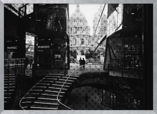 A framed, black and white photograph capturing a layered urban scene. The view, seen through a window with reflections of a curved, lit staircase, shows two people walking towards a large domed cathedral framed by modern glass buildings. Wall Art