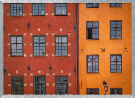 A framed close-up photograph of two adjacent, colorful building facades. The building on the left is a deep, rustic red, while the one on the right is a vibrant orange. Both feature numerous multi-paned windows with dark frames. Artwork