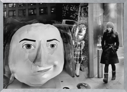 A surreal black and white photograph with a silver frame, featuring a giant, cartoonish head of a historical figure reflected in a window, juxtaposed with a young woman in a winter coat and beanie walking down a city sidewalk. Wall Art