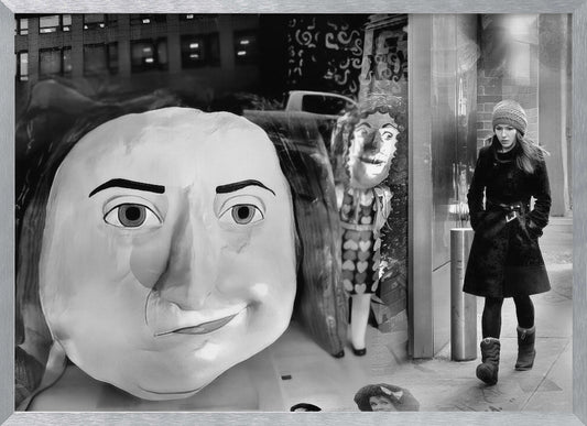 A surreal black and white photograph with a silver frame, featuring a giant, cartoonish head of a historical figure reflected in a window, juxtaposed with a young woman in a winter coat and beanie walking down a city sidewalk. Wall Art