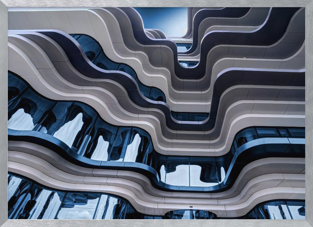 A low-angle view of a modern building with a striking, wavy facade. Alternating bands of light and dark grey create a fluid, ribbon-like pattern, with curved glass windows reflecting the deep blue sky. Artwork