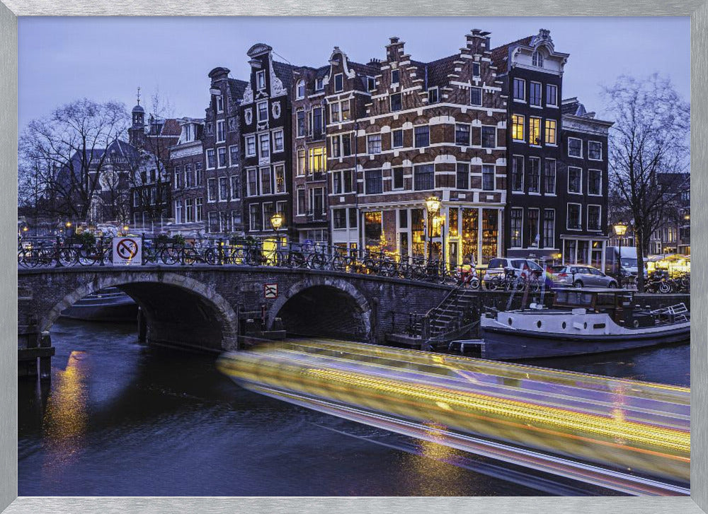 A long-exposure photograph of an Amsterdam canal at dusk, featuring a bright yellow light trail from a passing boat. In the background, illuminated historic canal houses line the water, and an arched stone bridge covered in bicycles crosses the canal. The entire image is presented in a silver frame. Print