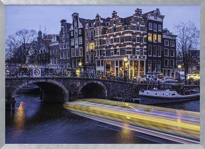 A long-exposure photograph of an Amsterdam canal at dusk, featuring a bright yellow light trail from a passing boat. In the background, illuminated historic canal houses line the water, and an arched stone bridge covered in bicycles crosses the canal. The entire image is presented in a silver frame. Print