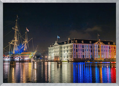 A vibrant nighttime photograph of the National Maritime Museum and a replica of the East Indiaman Amsterdam ship in a harbor. Both are illuminated with colorful lights that cast beautiful reflections on the dark water. Wall Art