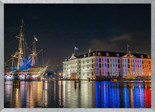 A vibrant nighttime photograph of the National Maritime Museum and a replica of the East Indiaman Amsterdam ship in a harbor. Both are illuminated with colorful lights that cast beautiful reflections on the dark water. Wall Art