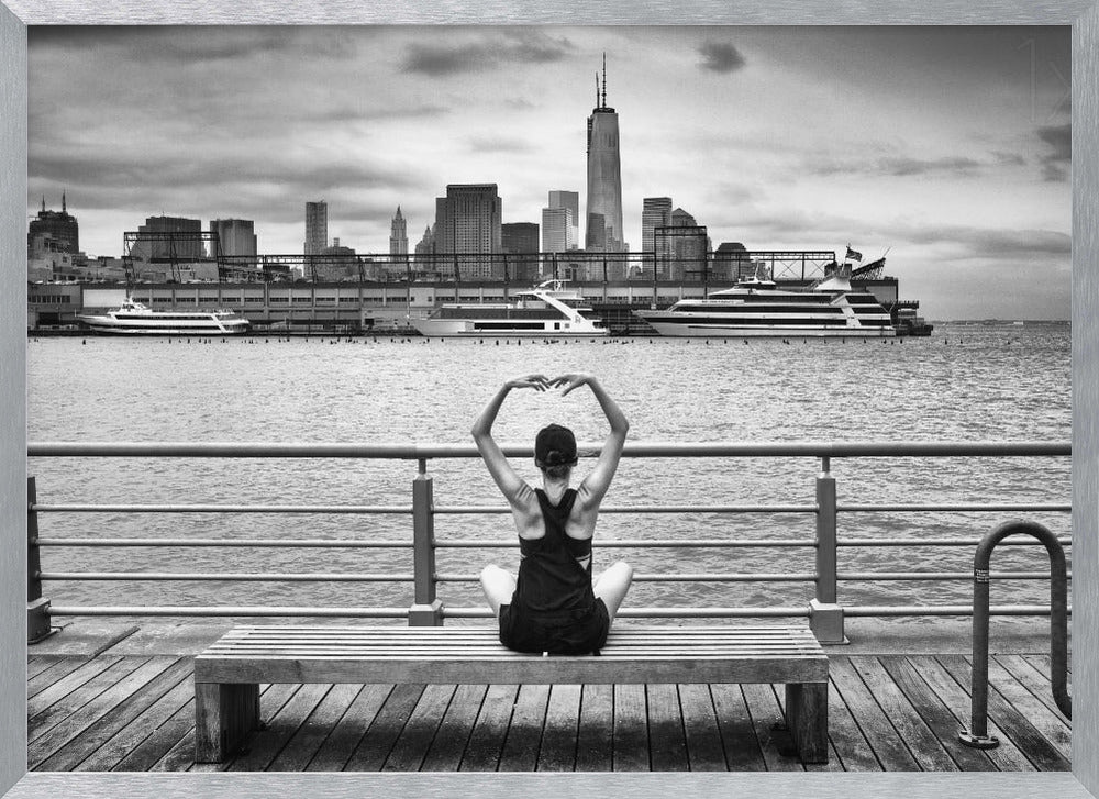 A framed black and white photograph showing the back of a woman sitting on a wooden bench on a pier. She is in a yoga pose with her arms raised, looking out at the Lower Manhattan skyline, including the Freedom Tower, across the water. Decor