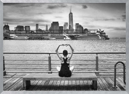 A framed black and white photograph showing the back of a woman sitting on a wooden bench on a pier. She is in a yoga pose with her arms raised, looking out at the Lower Manhattan skyline, including the Freedom Tower, across the water. Decor