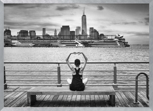 A framed black and white photograph showing the back of a woman sitting on a wooden bench on a pier. She is in a yoga pose with her arms raised, looking out at the Lower Manhattan skyline, including the Freedom Tower, across the water. Decor