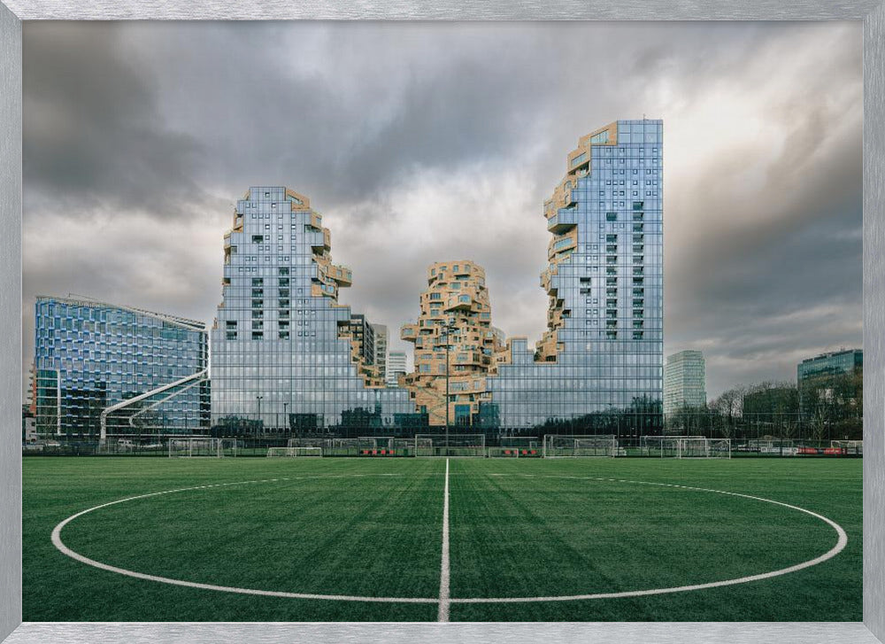 A wide-angle view from the center of a lush green soccer field looking towards a striking modern architectural complex with uniquely shaped glass and tan buildings under a dramatic, cloudy sky. Decor