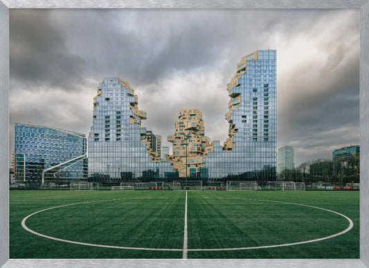 A wide-angle view from the center of a lush green soccer field looking towards a striking modern architectural complex with uniquely shaped glass and tan buildings under a dramatic, cloudy sky. Decor