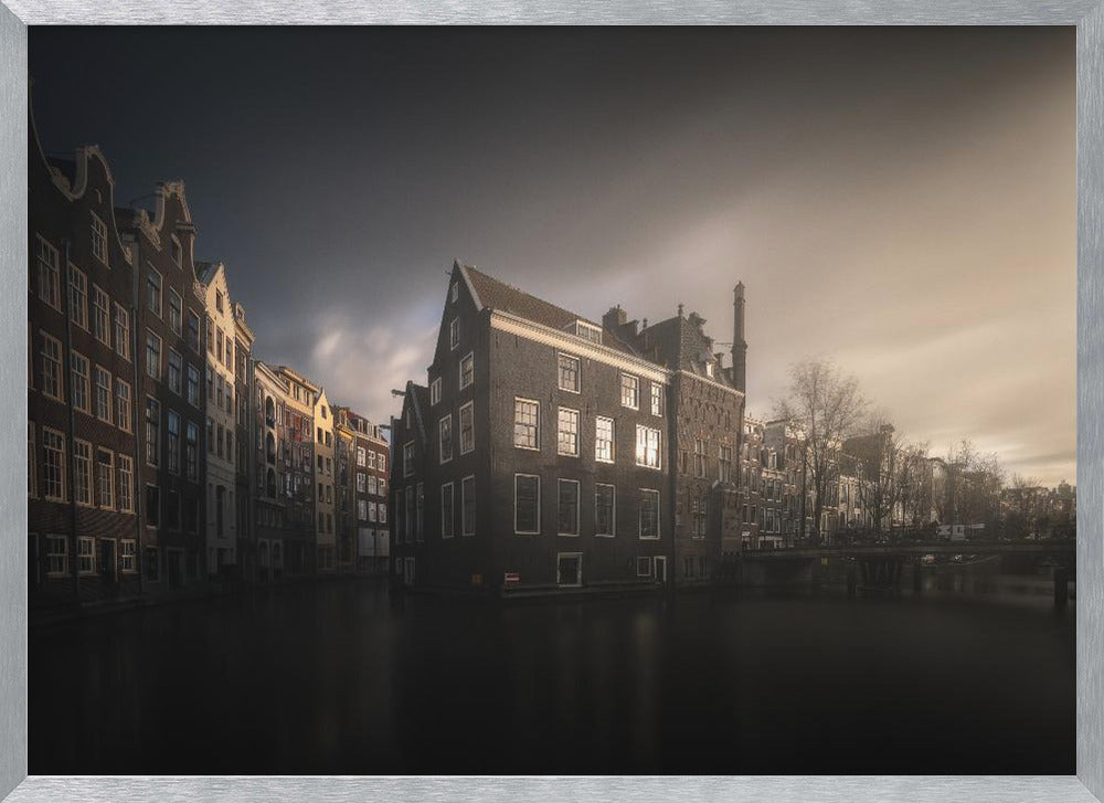 A moody, atmospheric photograph of historic canal houses in Amsterdam, viewed from the water. The scene is dark, with a dramatic, cloudy sky and a soft glow on the horizon, while some windows in the dark brick buildings are lit from within. The image is bordered by a silver frame. Wall Art