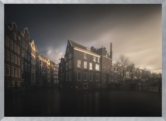 A moody, atmospheric photograph of historic canal houses in Amsterdam, viewed from the water. The scene is dark, with a dramatic, cloudy sky and a soft glow on the horizon, while some windows in the dark brick buildings are lit from within. The image is bordered by a silver frame. Wall Art
