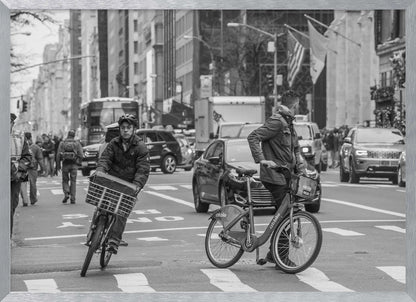 A black and white street photograph of a busy city intersection, framed in silver. In the foreground, two men are crossing with their bicycles amidst traffic, with one cyclist looking directly at the camera. Tall buildings and other pedestrians line the background, capturing a moment of everyday city life. Artwork